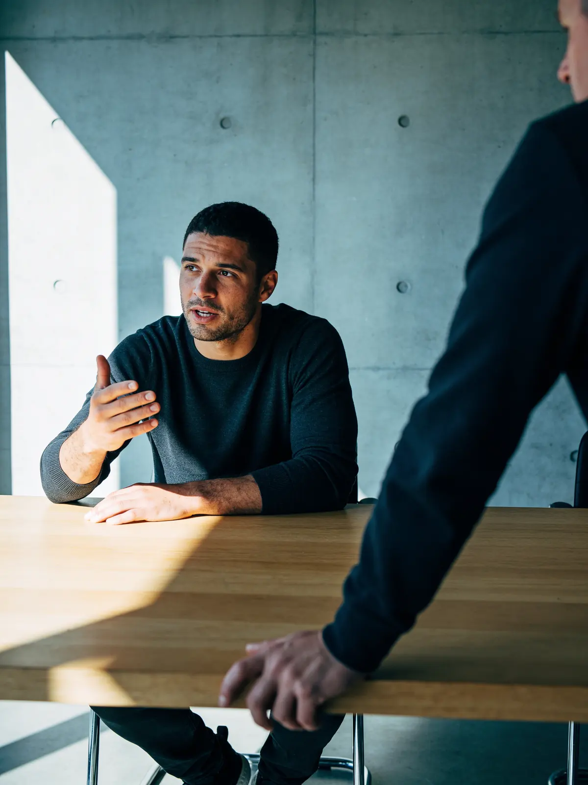 An athlete seated mid-conversation, hands on the table, an adviser's shoulder in the foreground.