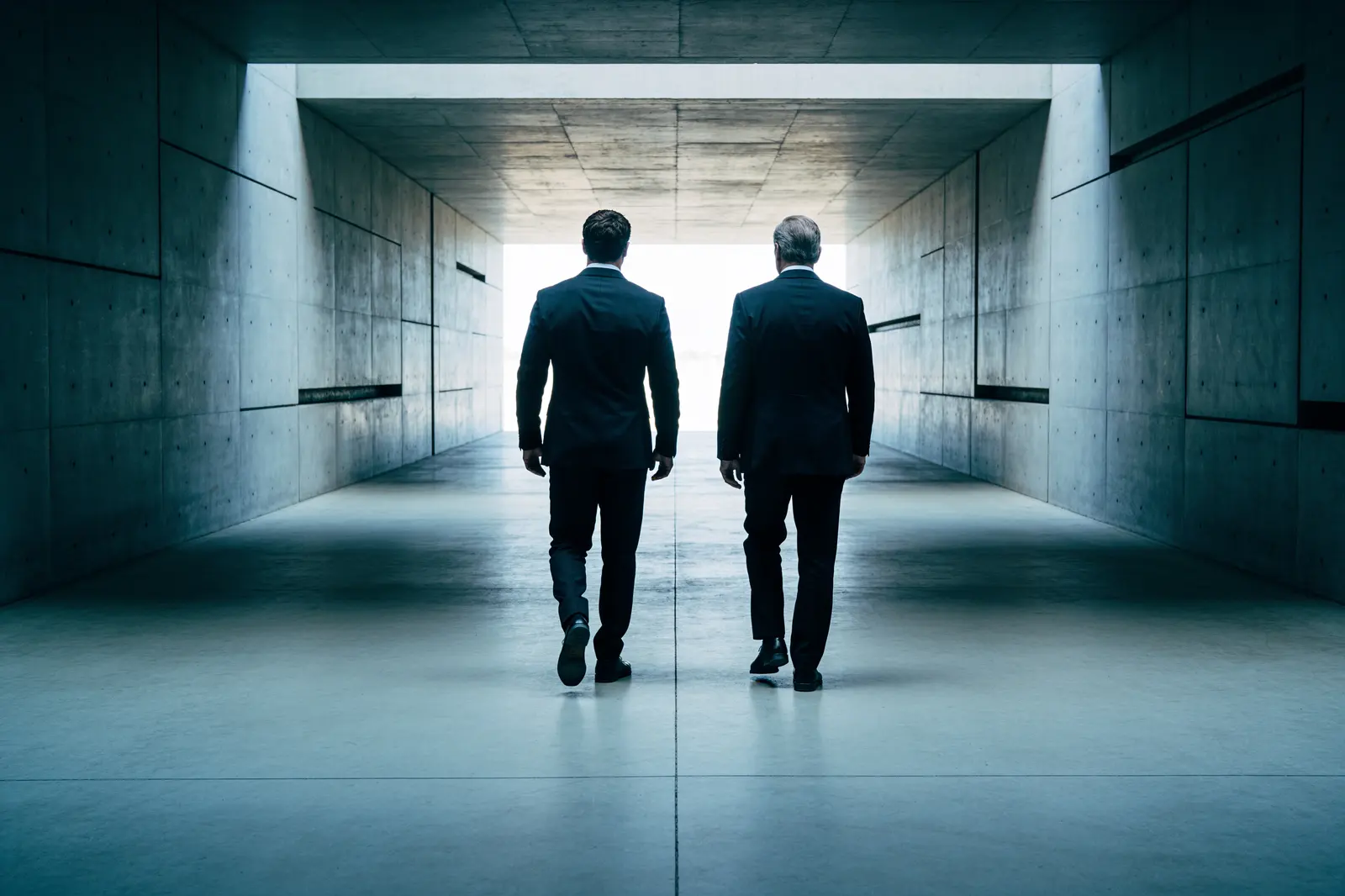 Two figures walking side by side down a concrete corridor toward the light.